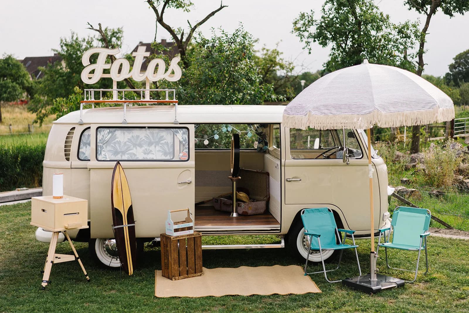 Cream-coloured photo booth bus with ‘Fotos’ sign, parasol, surfboard, printer and folding chairs in a garden