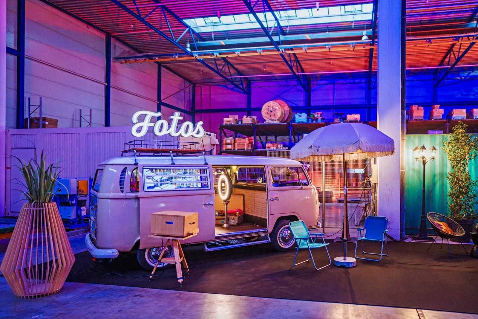 Der Fotobulli mit Leuchtschrift „Fotos“, Sonnenschirm und Klappstühlen in einer bunt beleuchteten Eventhalle