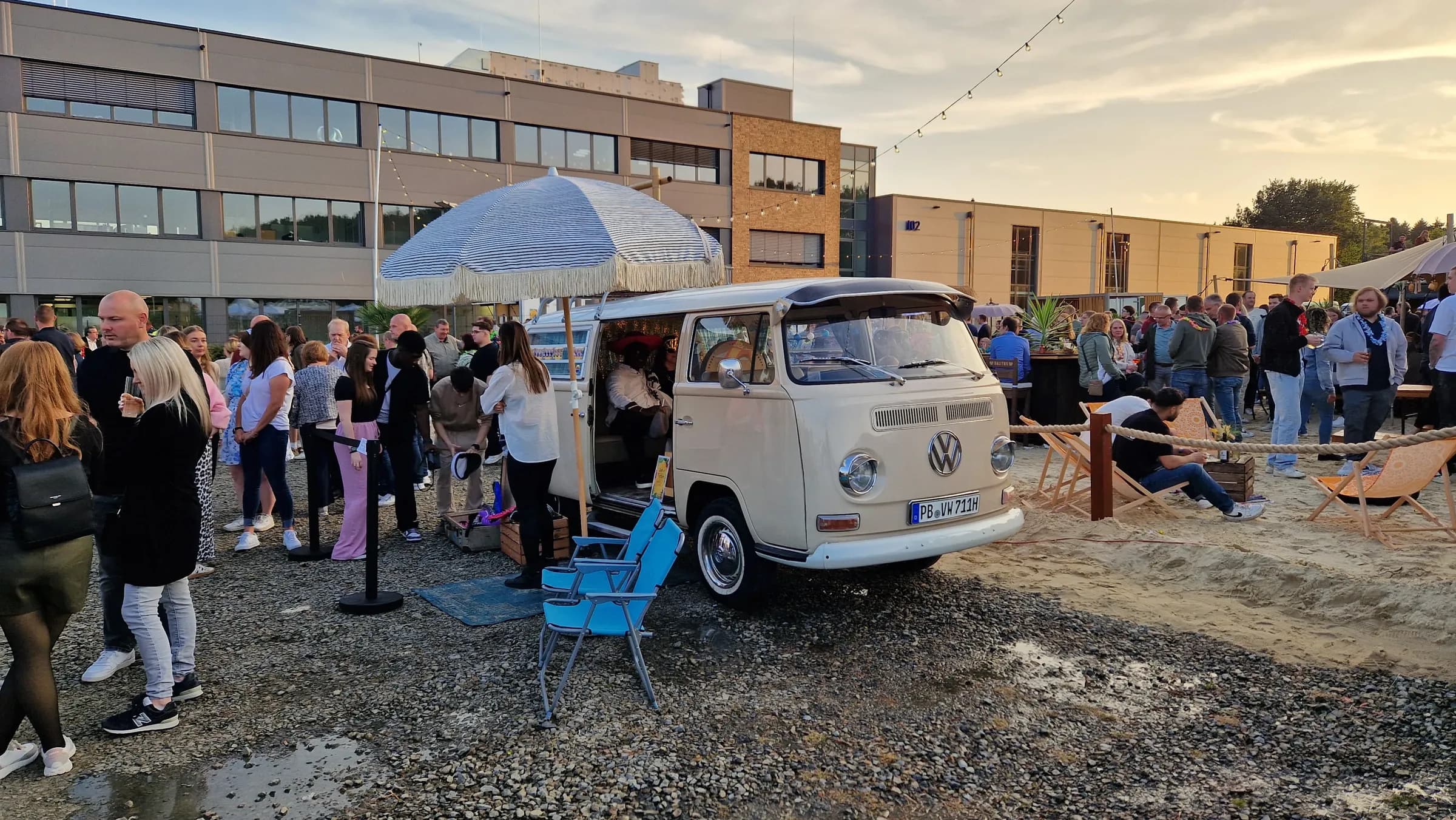 Cream-coloured photo booth bus with a fringed parasol at a company party, with guests queuing in front and deckchairs in the sand
