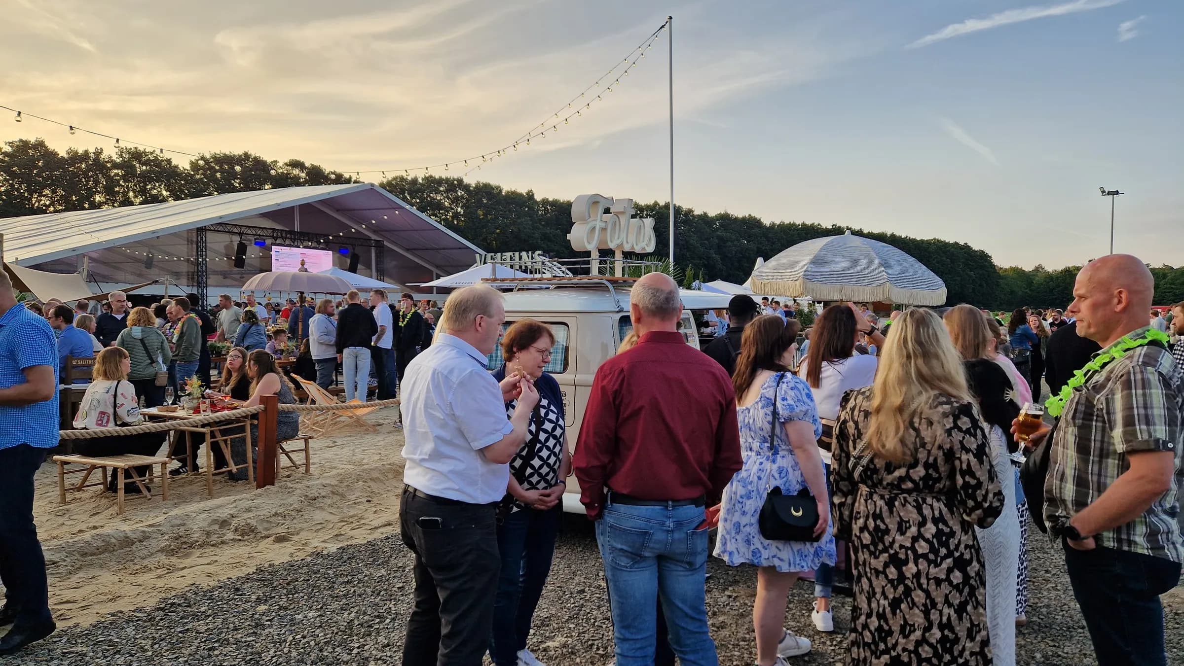 Lots of guests standing around the photo booth bus with its illuminated ‘Fotos’ sign in the evening, with a stage tent and beach area in the background
