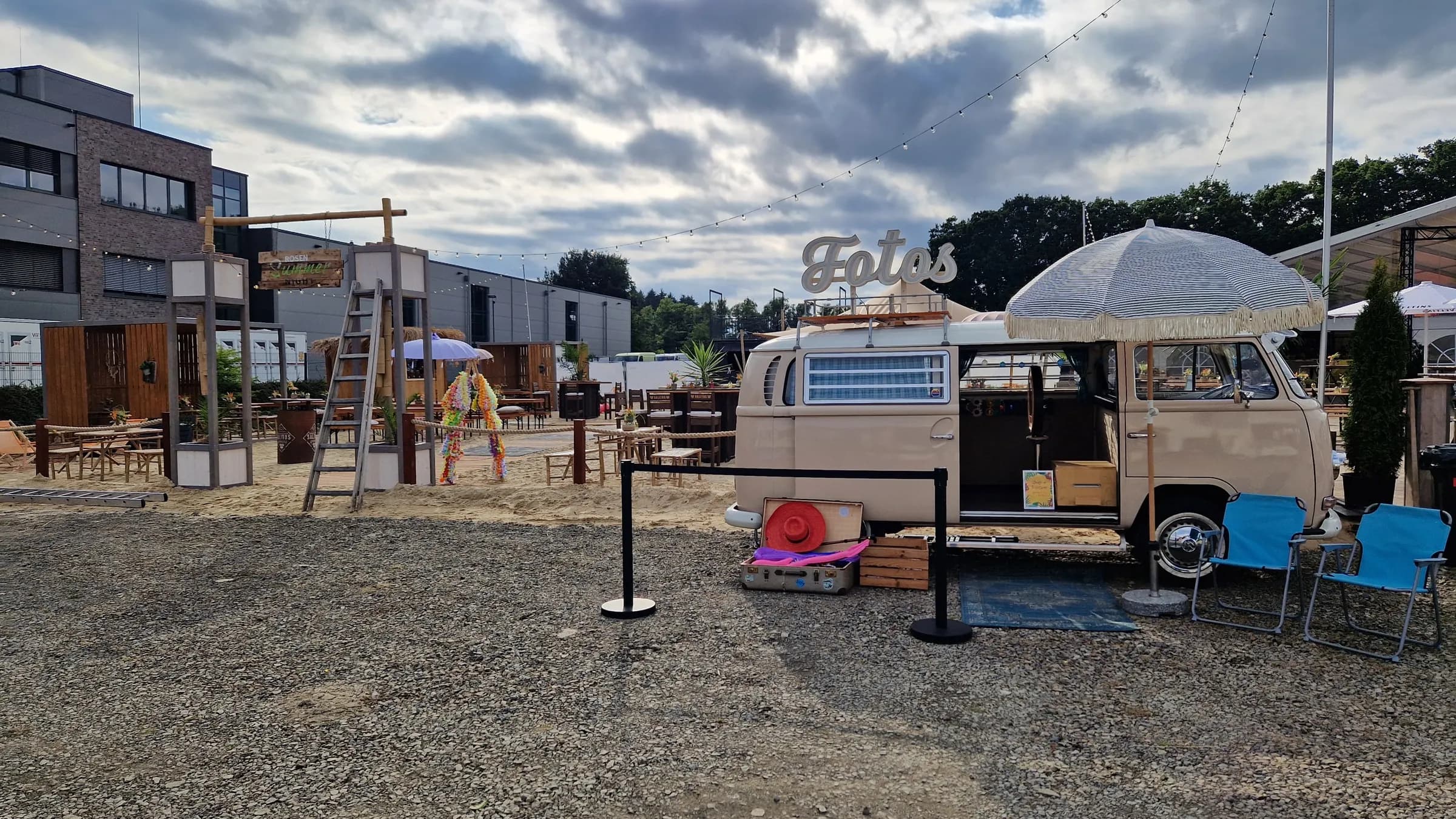 Photo booth bus with ‘Fotos’ neon sign and a parasol next to a beach bar backdrop on a company site