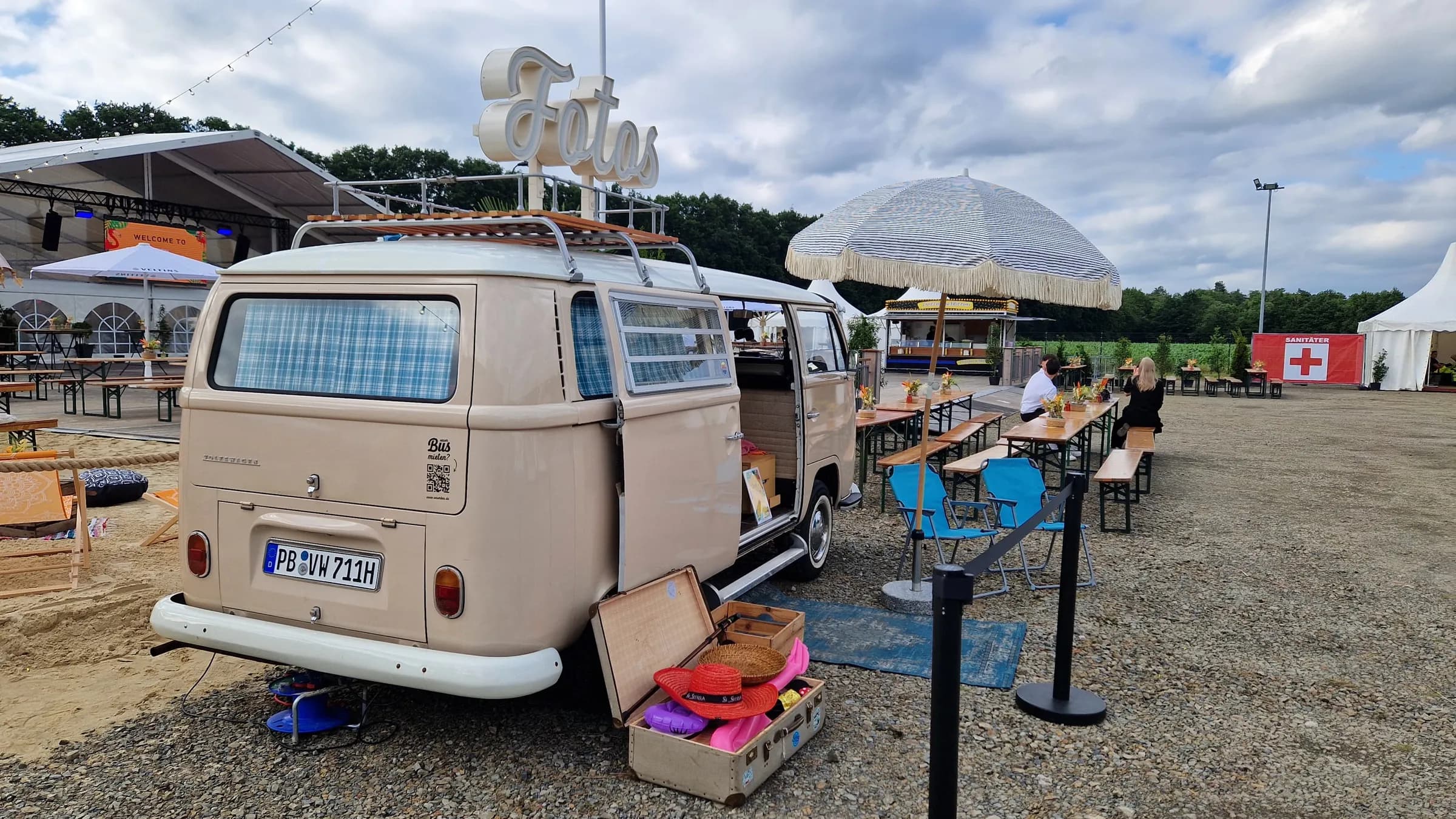 Photo booth bus seen from behind on a gravel area, with an open suitcase full of props in front of it, next to a parasol and beer tables
