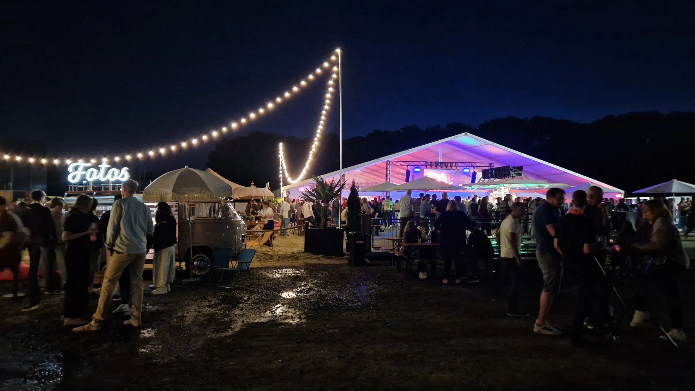 Fotobulli mit Leuchtschrift „Fotos“ unter Lichterketten am Abend, daneben ein beleuchtetes Festzelt voller Gäste