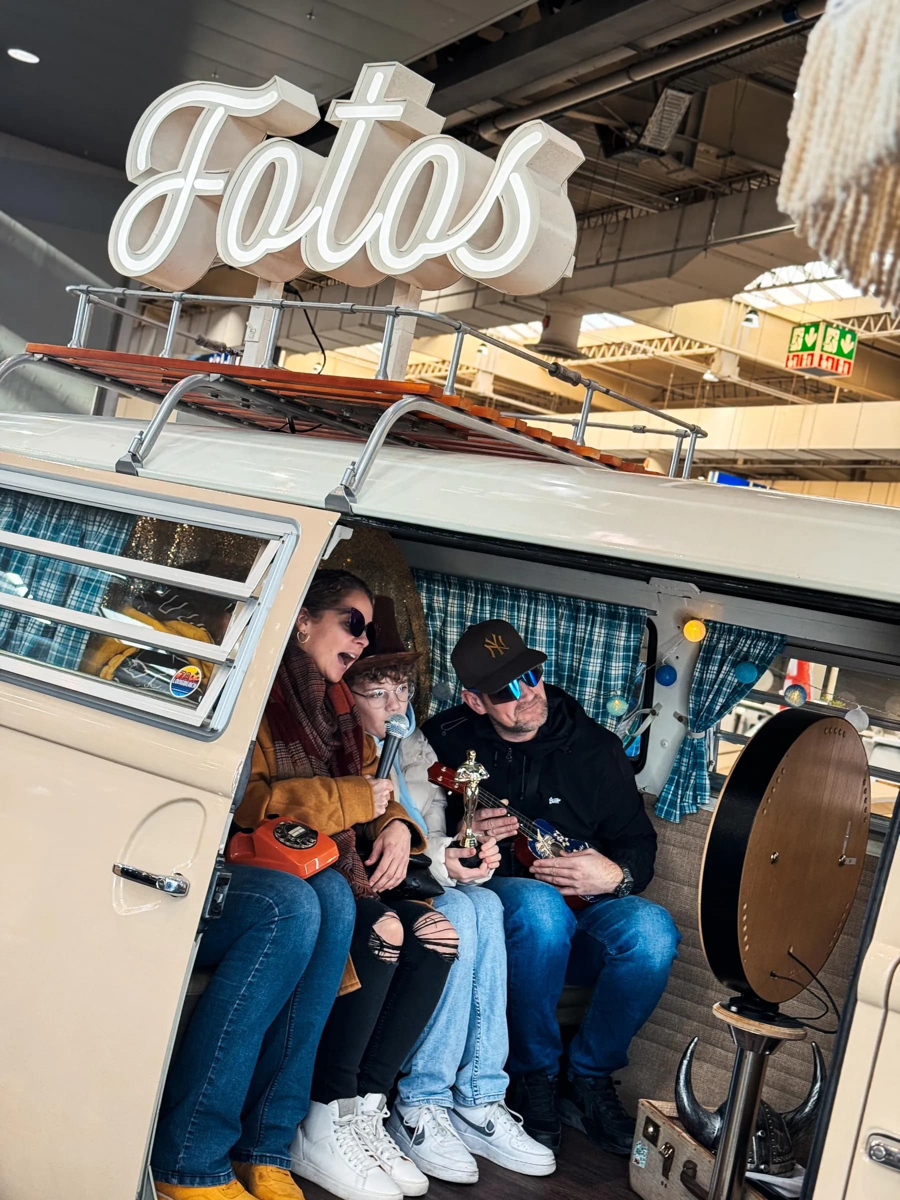 Drei Gäste mit Mikrofon, Ukulele und Trophäe im Fotobulli auf der ABF Hannover, auf dem Dach die Leuchtschrift „Fotos“