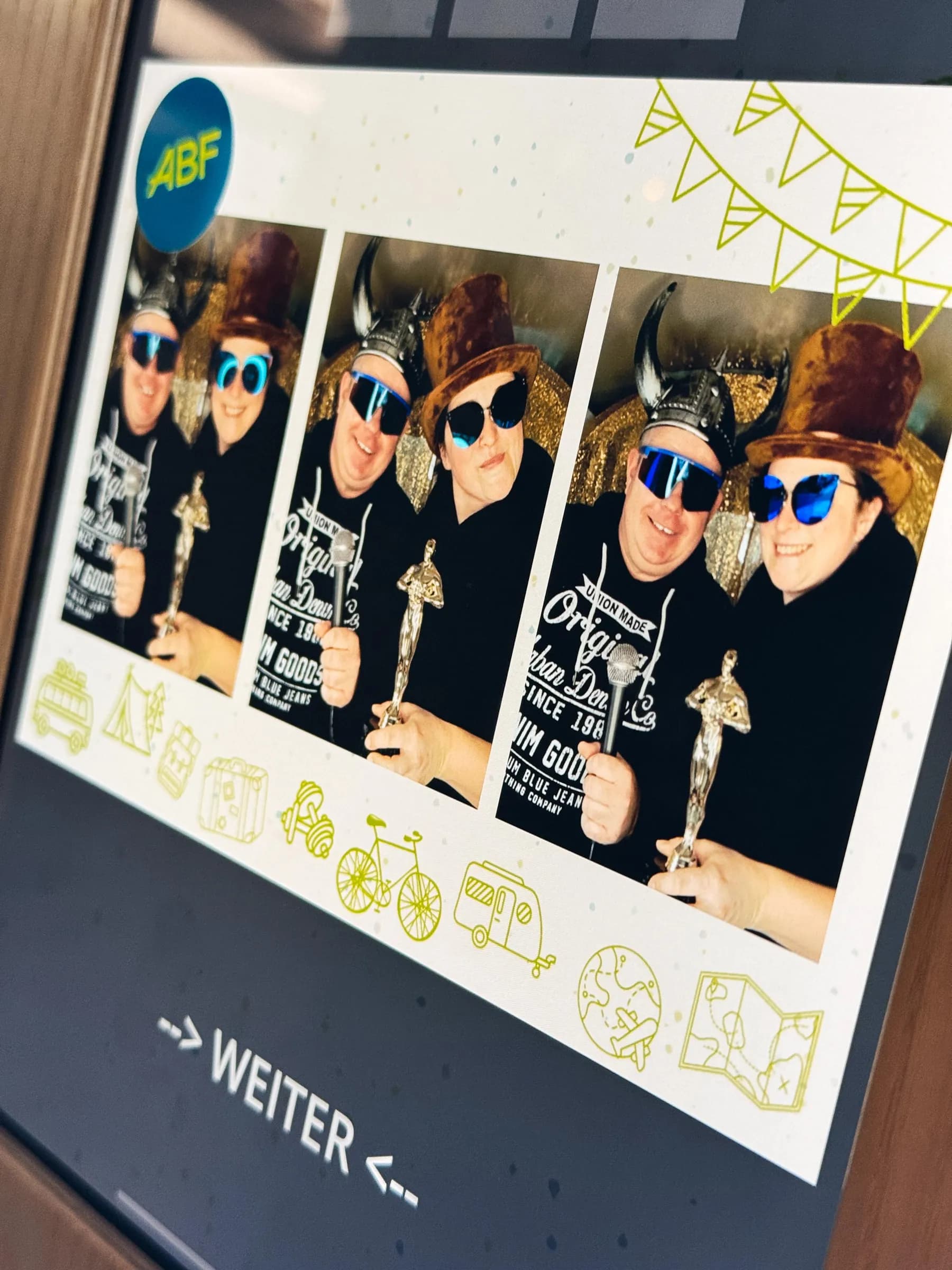 Display im Fotobulli mit ABF-Fotolayout: zwei Gäste mit Wikingerhelm, Zylinder, Sonnenbrillen und goldener Trophäe