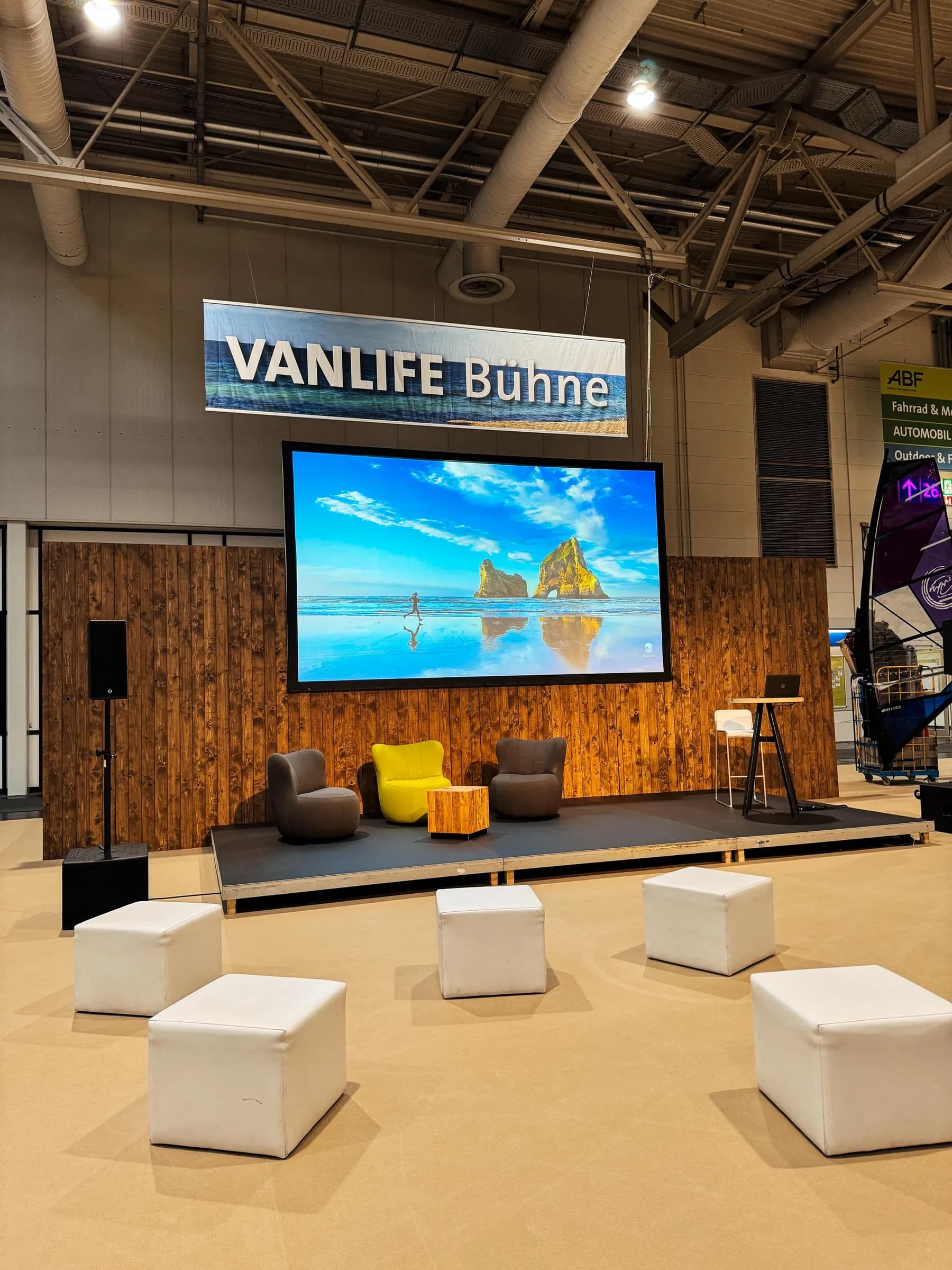 Vanlife-Bühne auf der ABF in Hannover mit großer Leinwand, Sesseln, Stehtisch und weißen Sitzwürfeln davor