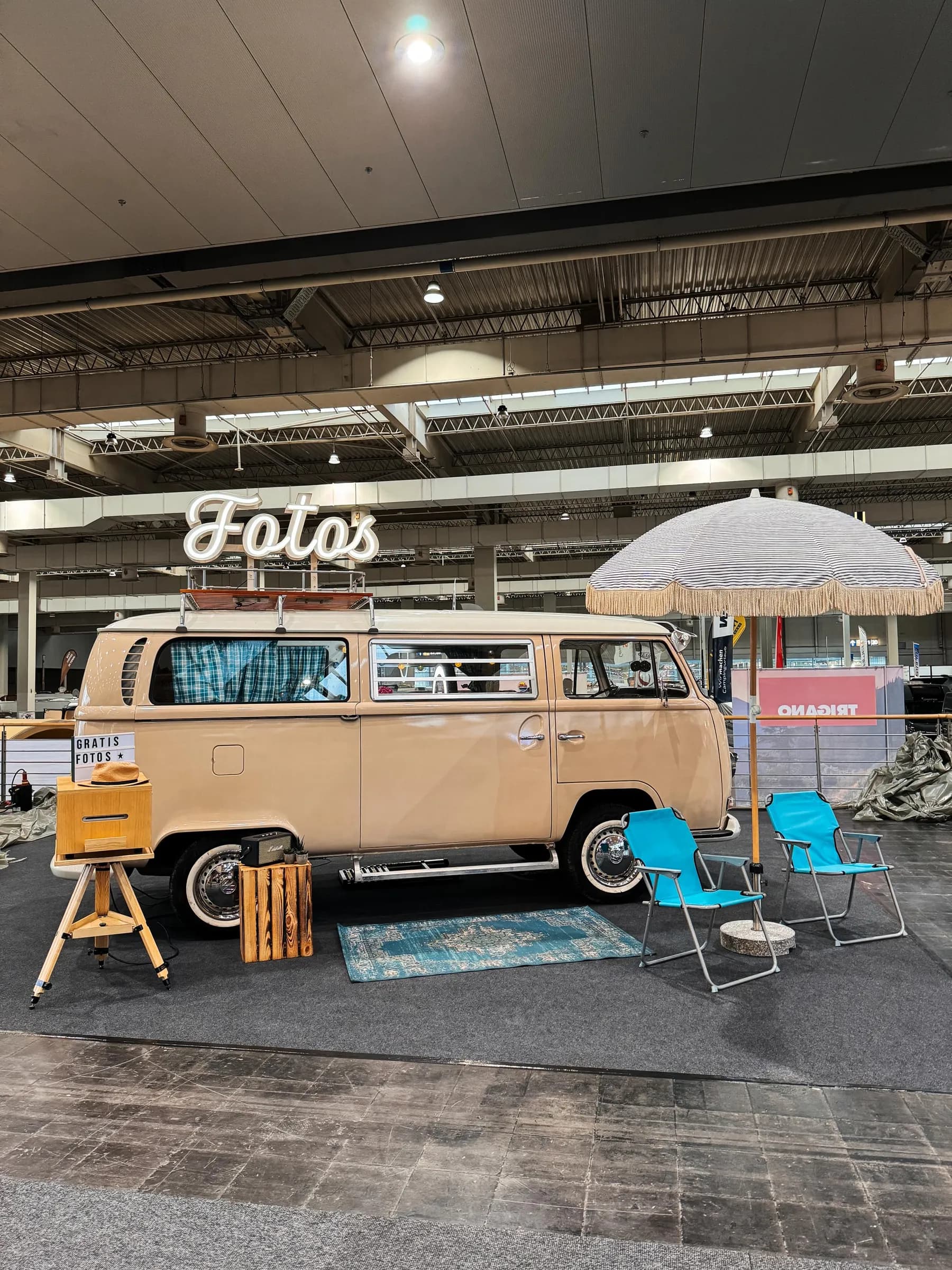 Beiger Fotobulli mit Leuchtschrift „Fotos“, Sonnenschirm, Teppich und blauen Klappstühlen in einer Messehalle der ABF