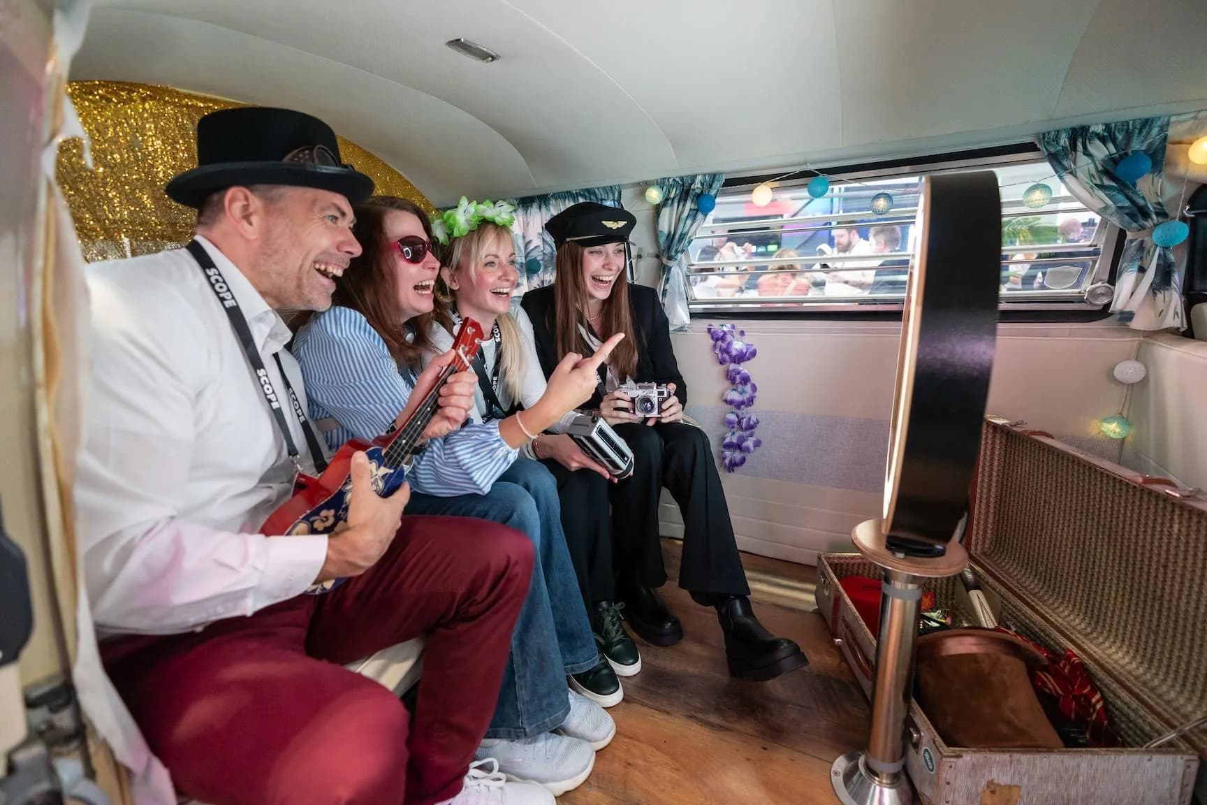Vier lachende Gäste mit Hut, Ukulele und Kamera posieren im Fotobulli vor der Fotobox, daneben ein Koffer mit Props