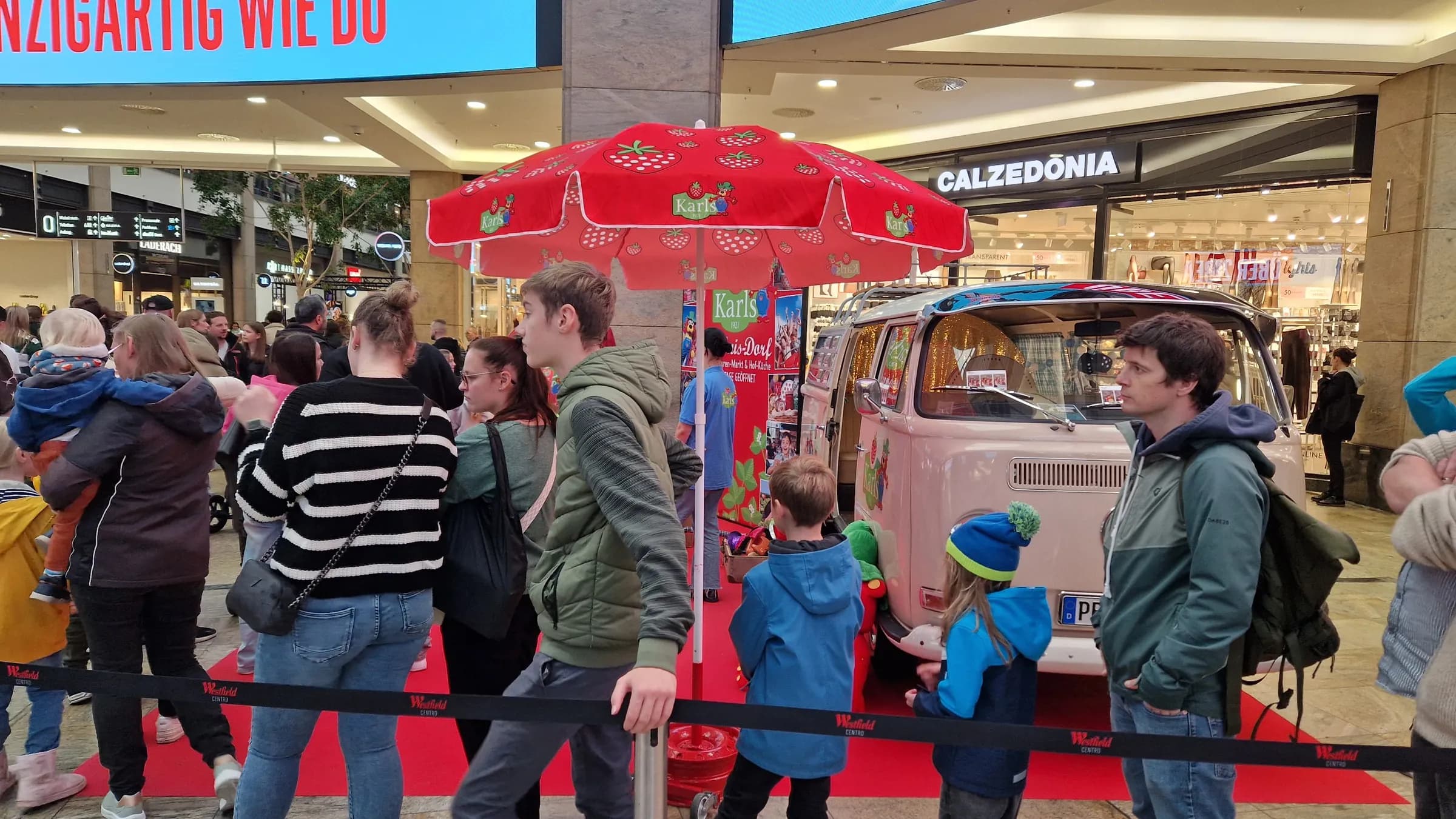 Families and children standing at the barrier in front of the photo booth bus under a red strawberry parasol in the shopping centre
