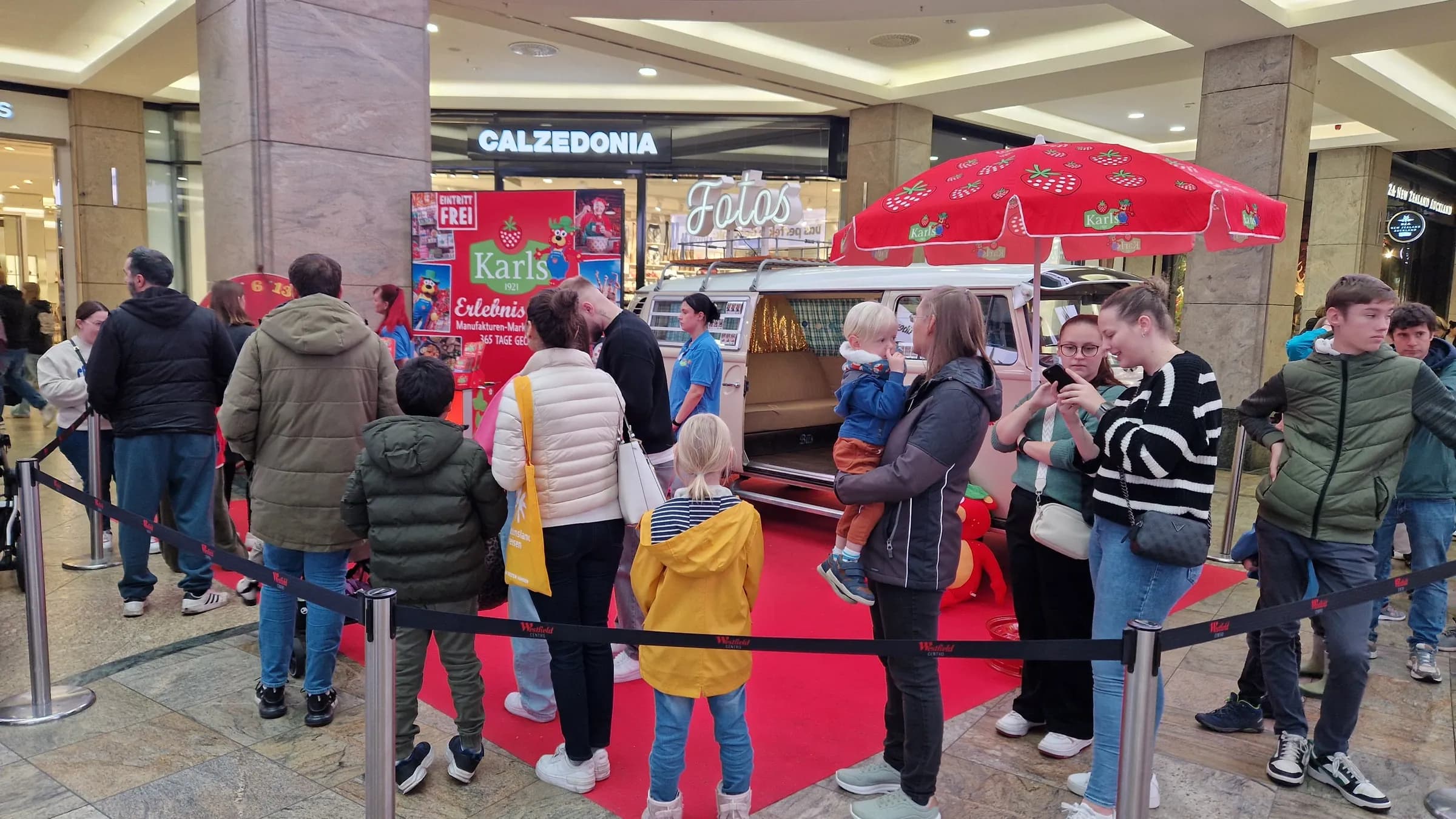 Photo booth bus with its sliding door open, a Karls parasol and an illuminated ‘Fotos’ sign in a shopping centre, with families waiting in front