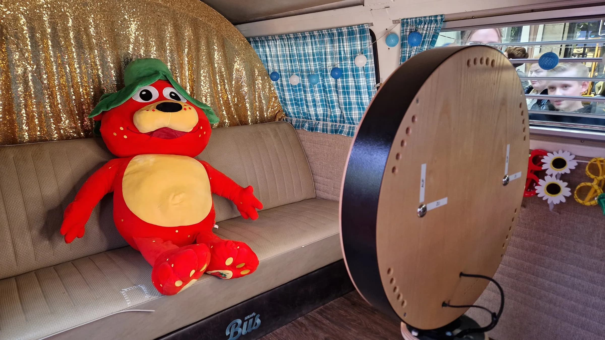 Red Karls plush mascot on the bench seat in the photo booth bus, in front of a gold glitter curtain and opposite the photo booth