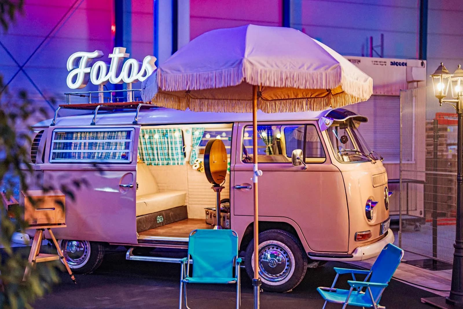 Photo booth bus with ‘Fotos’ neon sign, checked curtains, bench seat and photo booth inside, with a fringed parasol and folding chairs in front