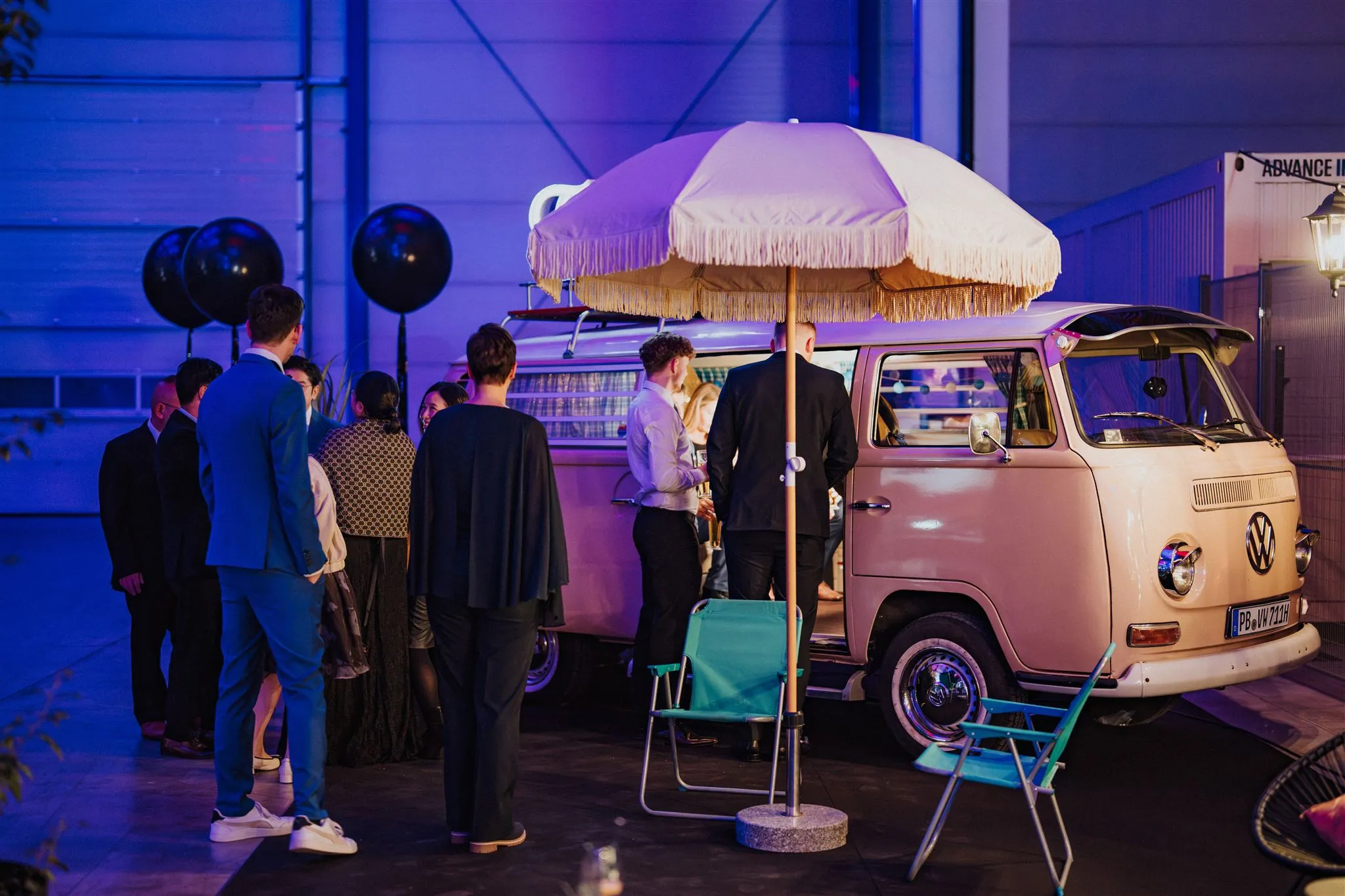 Guests in evening wear stand by the photo booth bus under a fringed parasol, waiting at the open sliding door for their photo