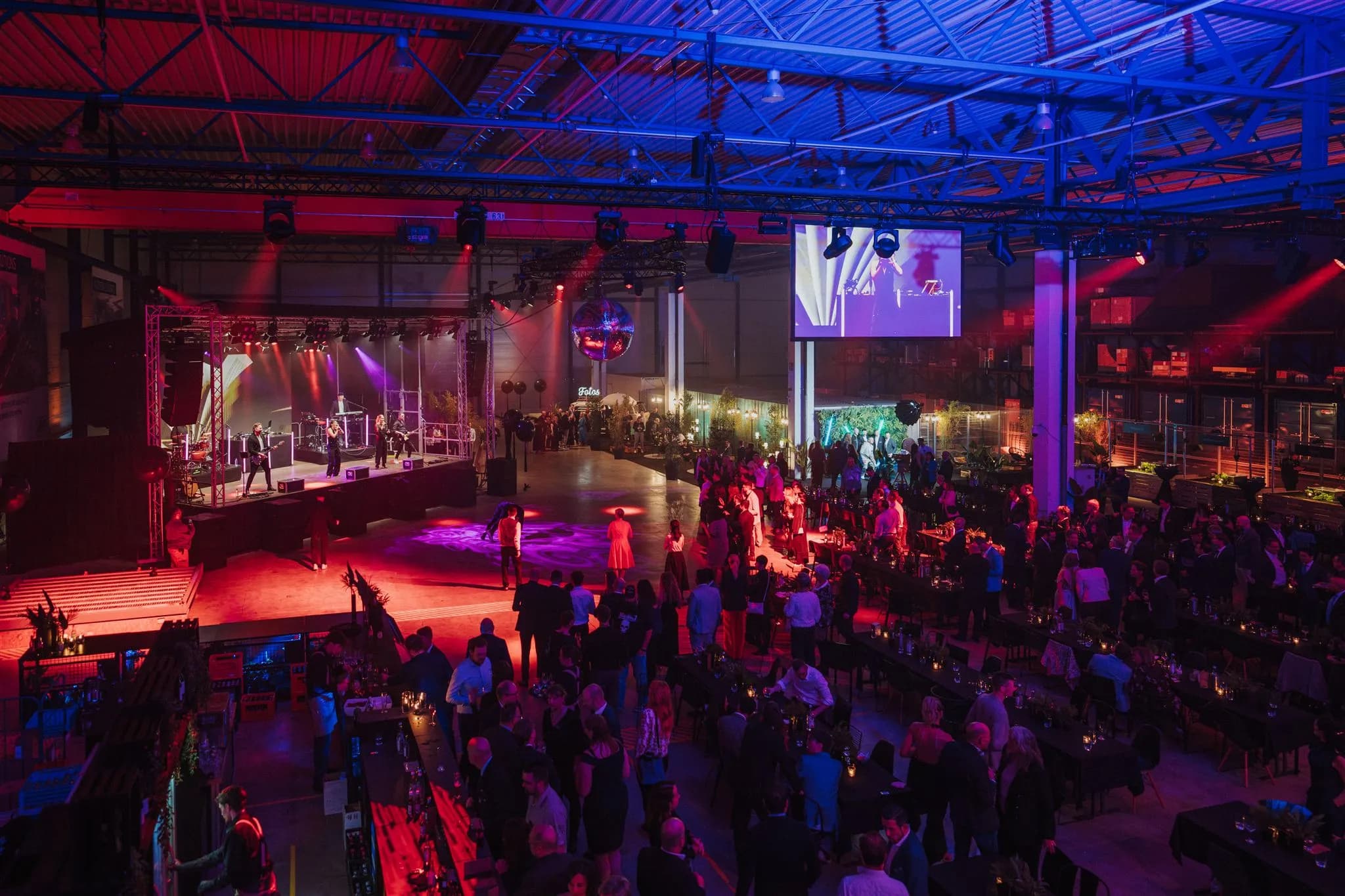 Company party in a hall with a live band on stage, guests at long tables and the photo booth bus with an illuminated sign in the background