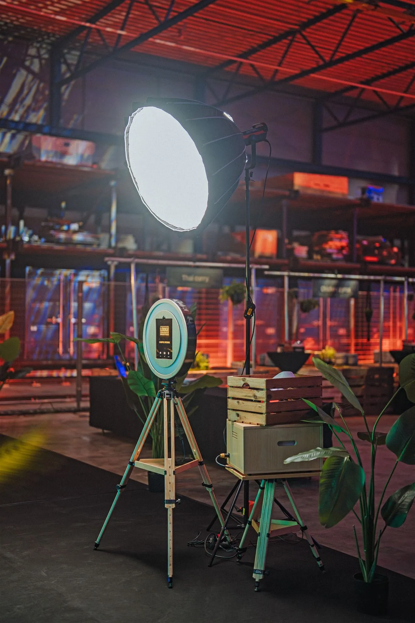 Photo booth with a round ring light and display on a wooden stand, next to it a printer in a wooden box and a softbox in an illuminated hall
