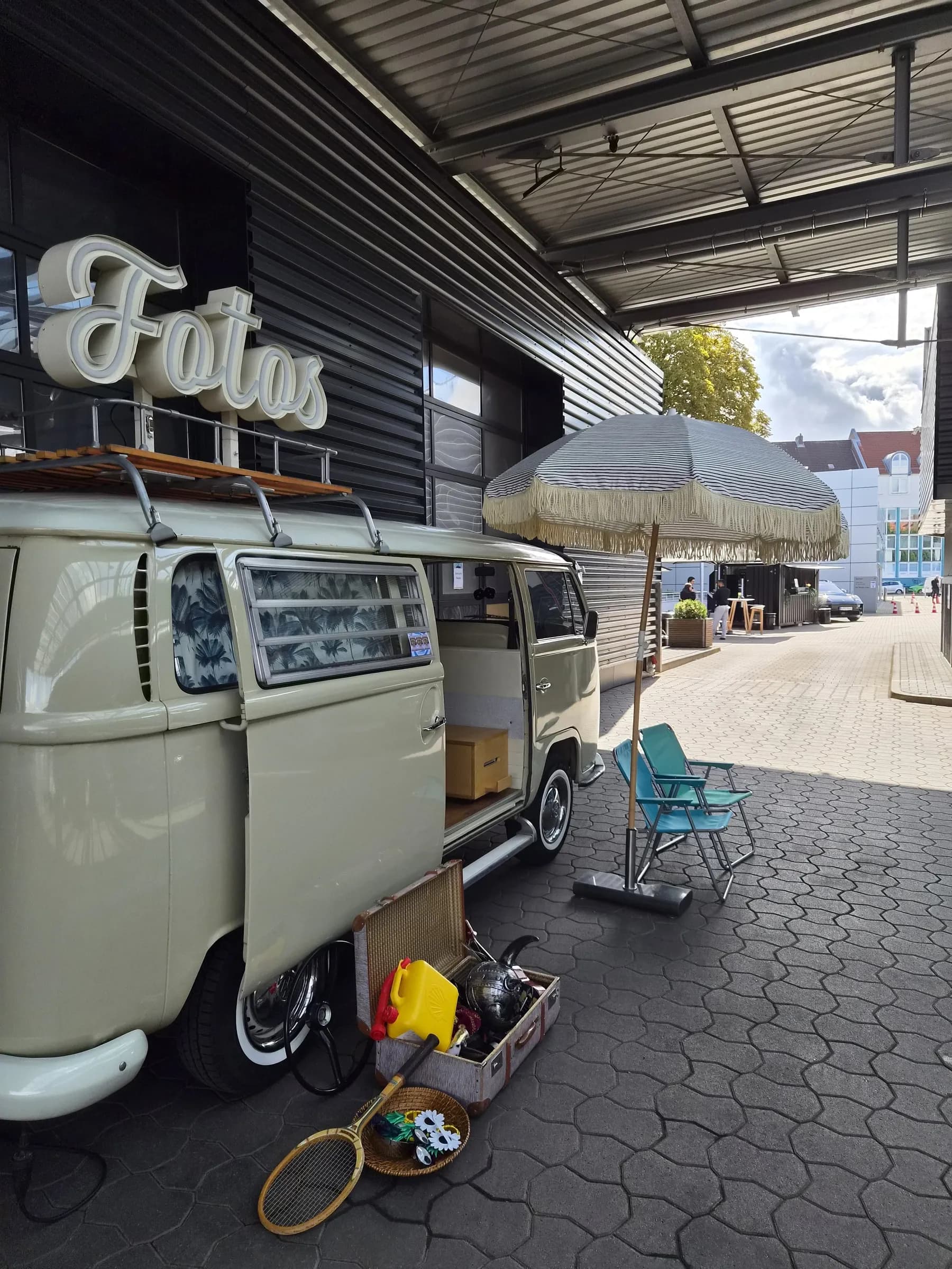 Photo booth bus with an illuminated ‘Fotos’ sign on the roof in front of Porsche Zentrum Hannover, with a parasol, chairs and props beside it