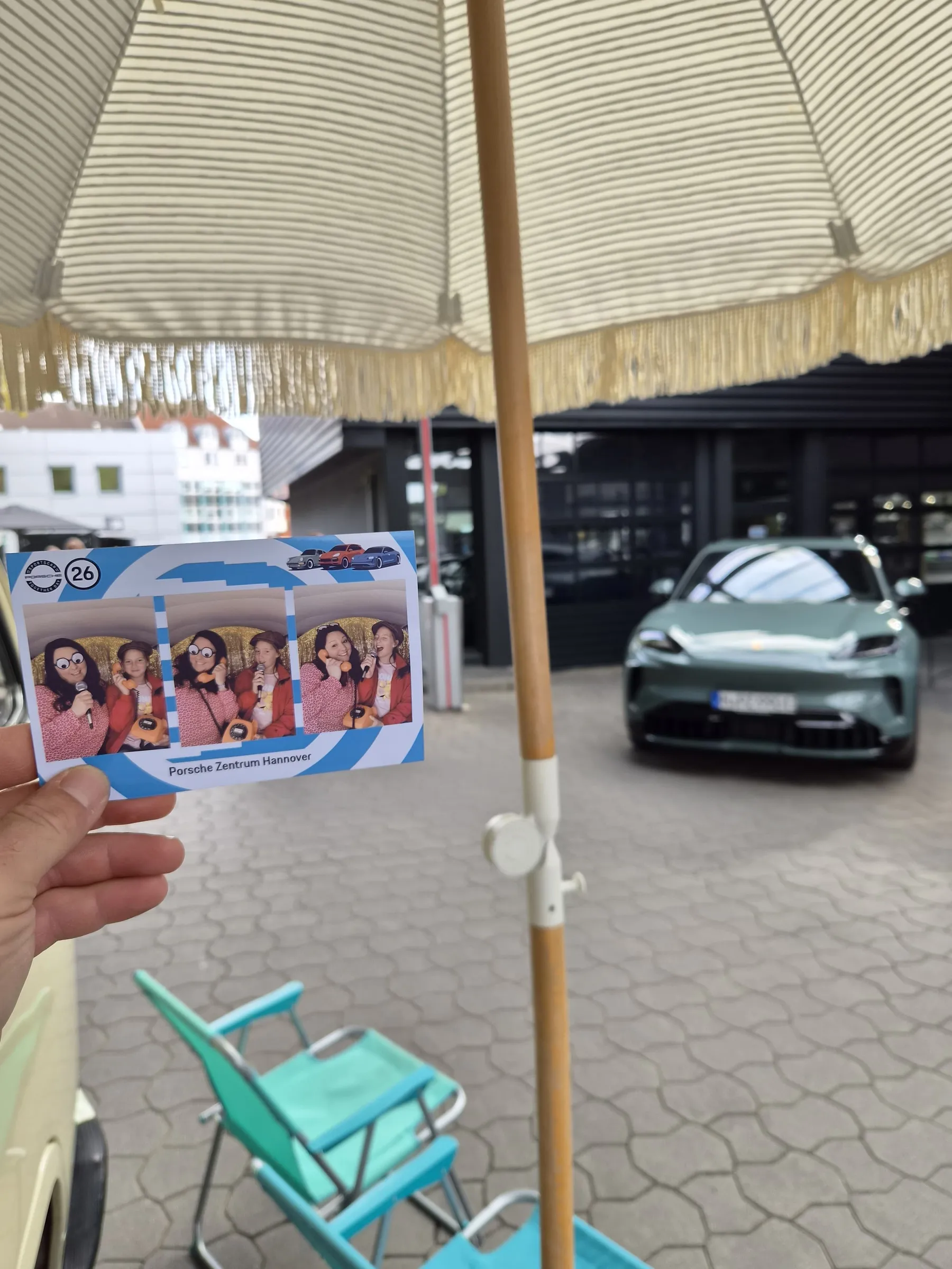 Hand holding a photo strip with a Porsche Zentrum Hannover layout, with a parasol, deckchair and green Porsche in the background