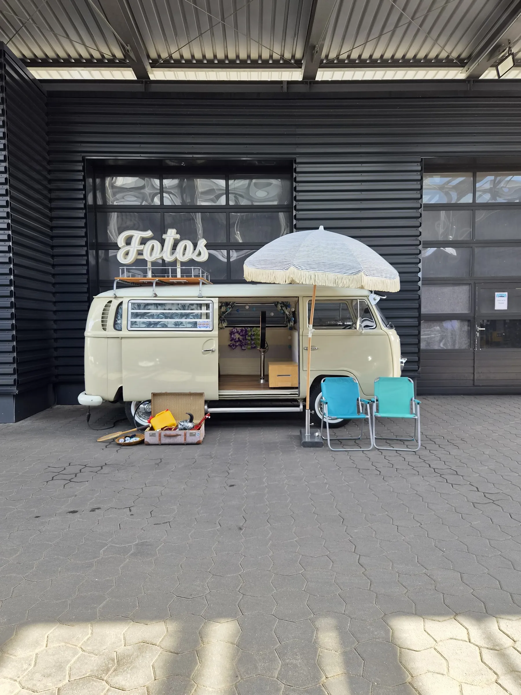 Photo booth bus with open sliding door, illuminated ‘Fotos’ sign, parasol, folding chairs and props case in front of a dark hall