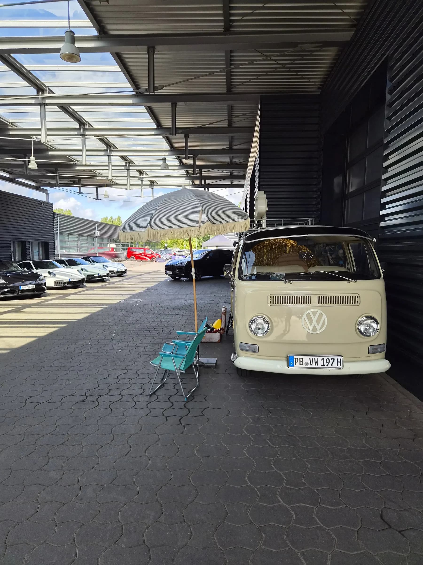 Beige VW Bulli with a parasol and folding chairs under a canopy, with several Porsches parked alongside