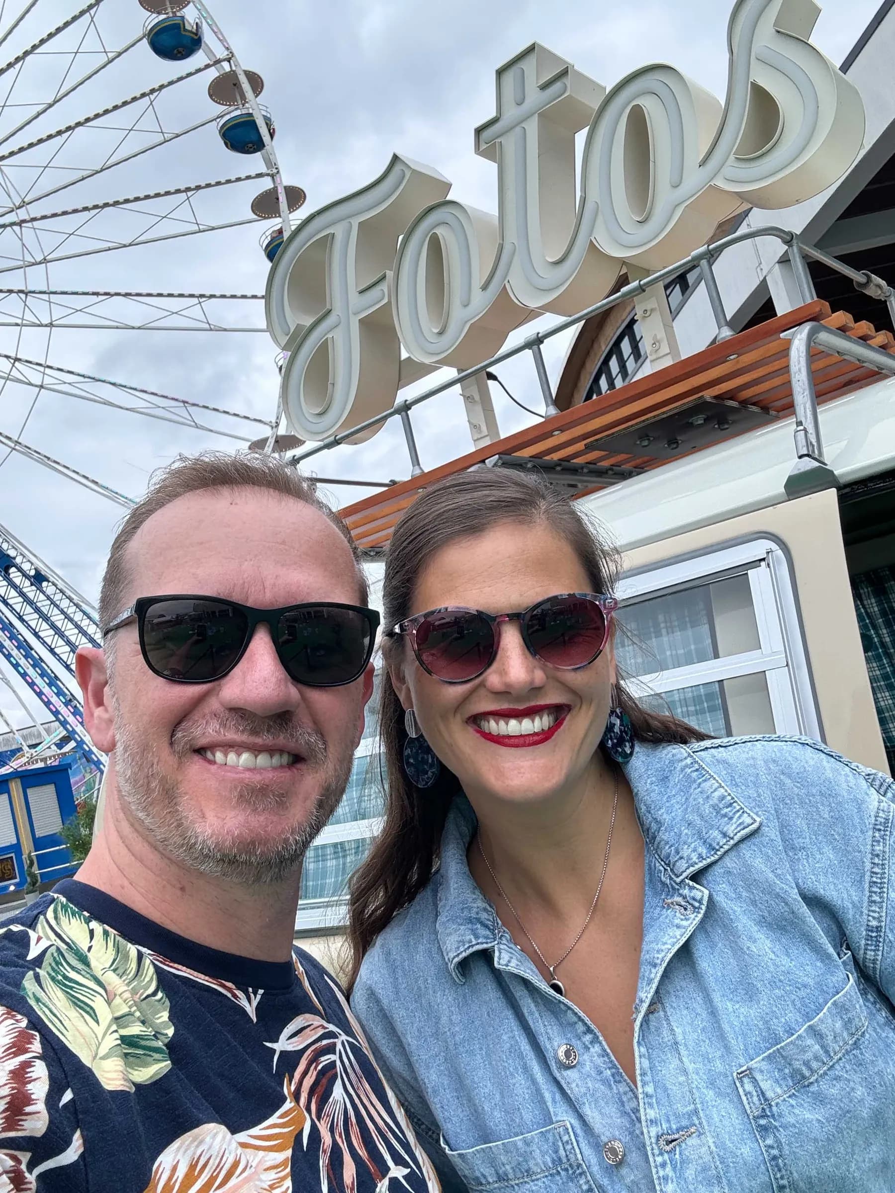 Mann und Frau mit Sonnenbrillen machen ein Selfie vor dem Fotobulli mit Fotos-Leuchtschrift und Riesenrad