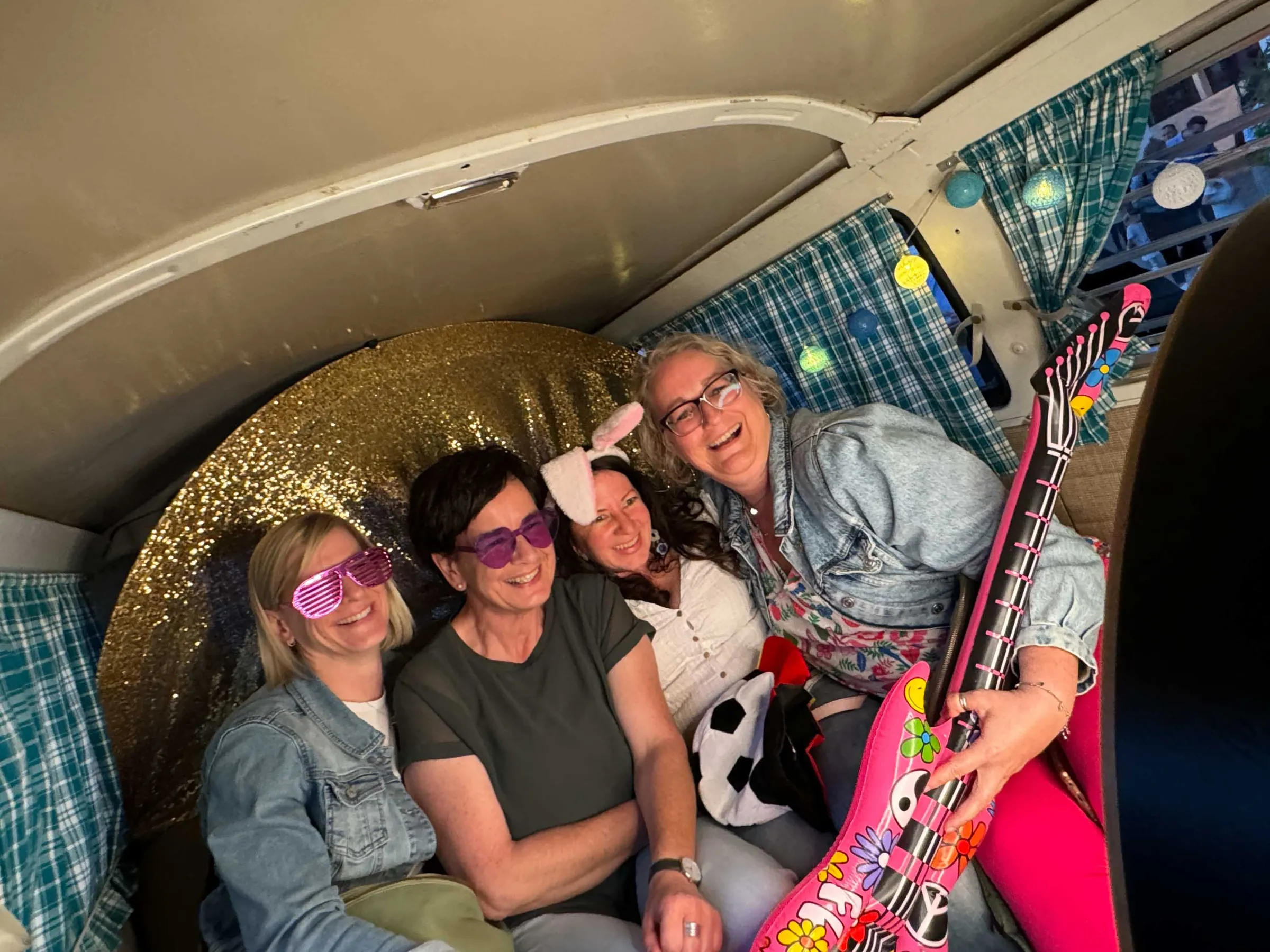 Vier lachende Frauen mit Partybrillen, Hasenohren und pinker Luftgitarre im Fotobulli vor glitzernder Goldwand
