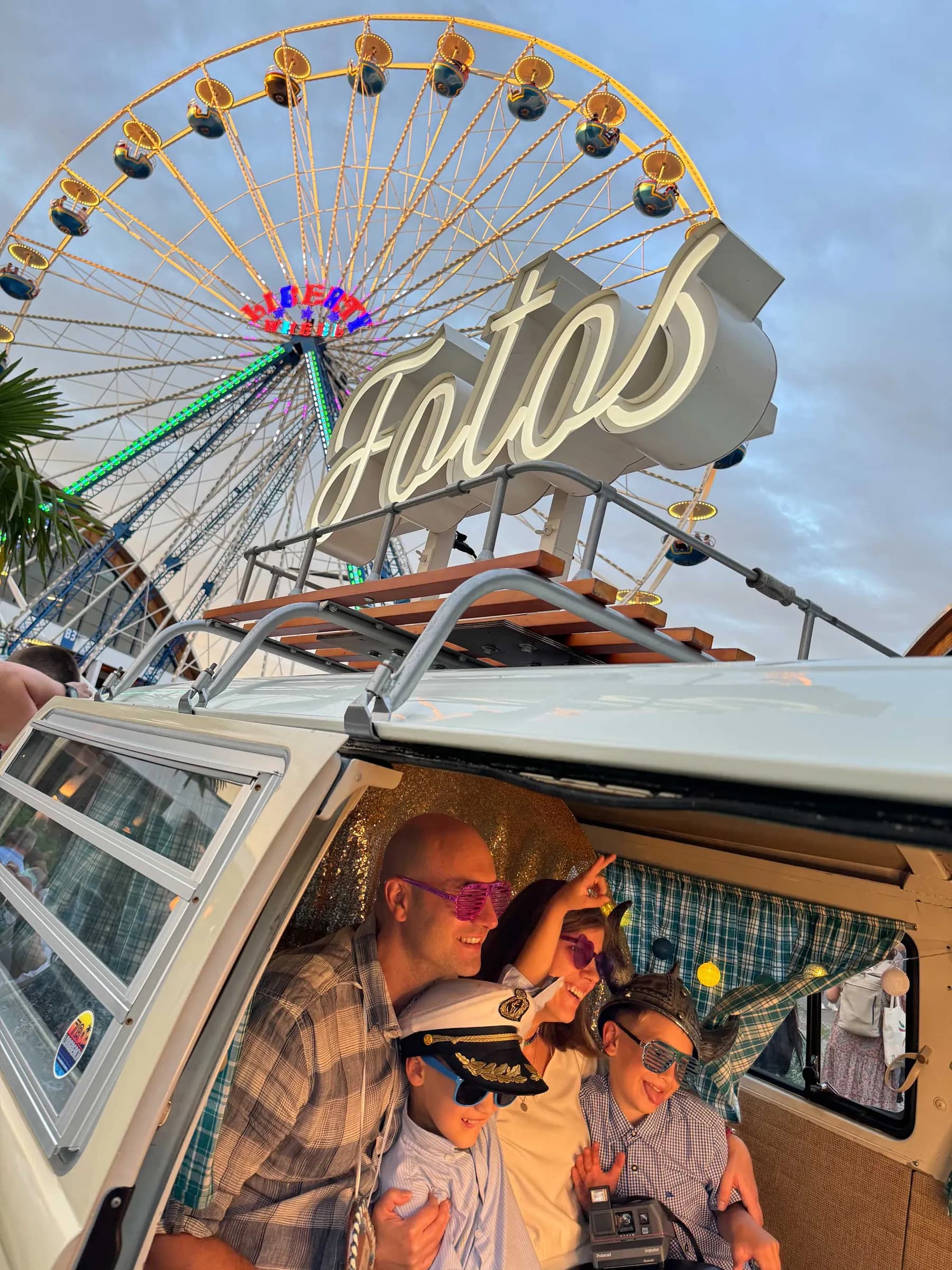 Familie mit Kapitänsmütze und Partybrillen posiert im Fotobulli, darüber Leuchtschrift „Fotos“ und ein Riesenrad