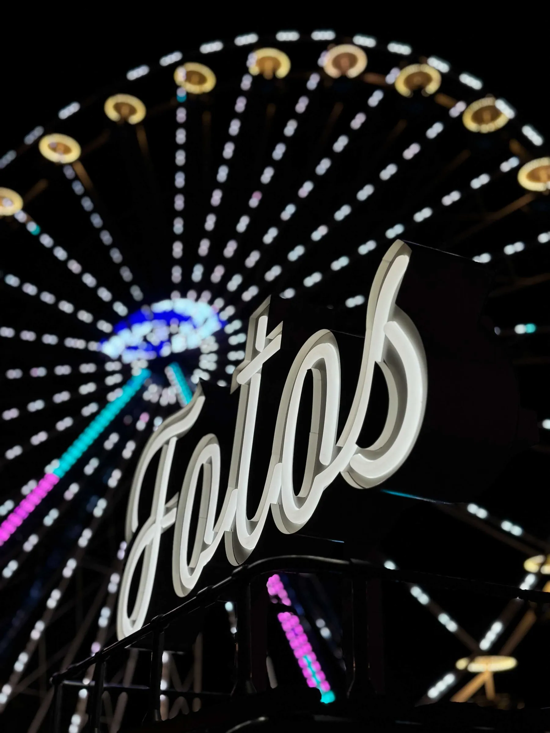 Leuchtschrift „Fotos“ auf dem Dach des Fotobullis bei Nacht, dahinter ein beleuchtetes Riesenrad