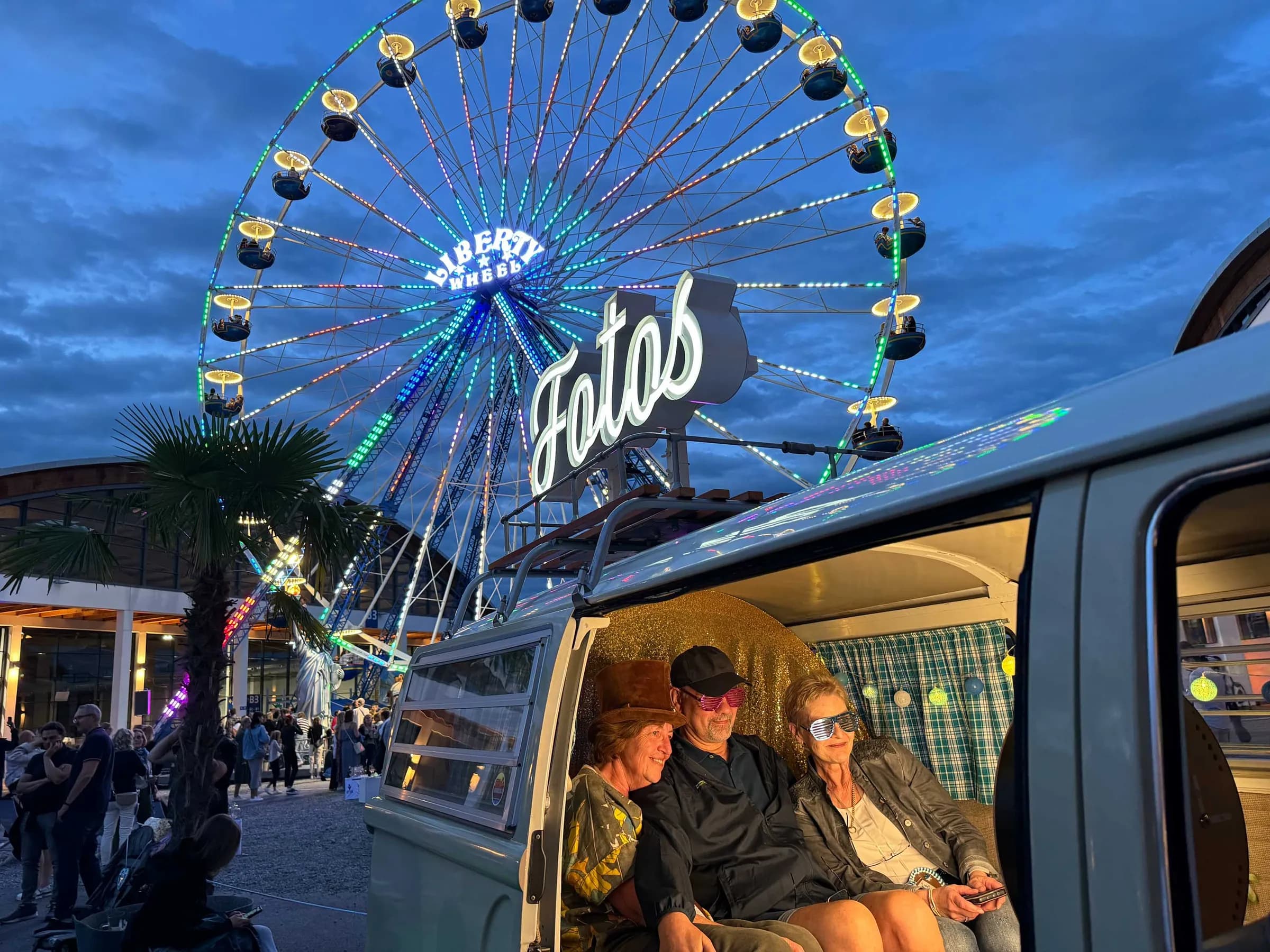 Drei Gäste mit Hut und Partybrillen im Fotobulli, dahinter Fotos-Leuchtschrift und beleuchtetes Riesenrad am Abend
