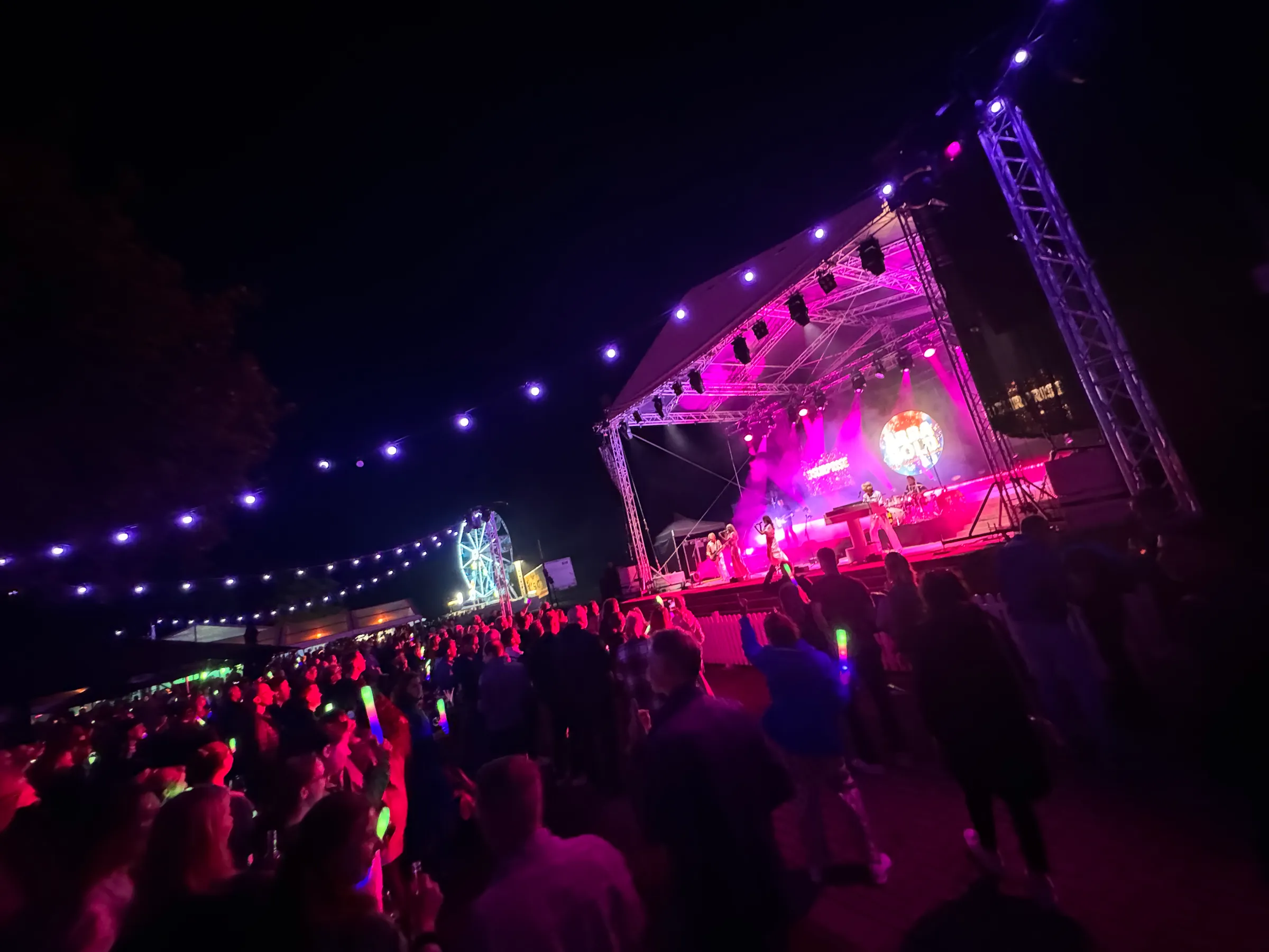 Night-time summer party with a band on a pink-lit stage, audience holding glow sticks, fairy lights and a Ferris wheel
