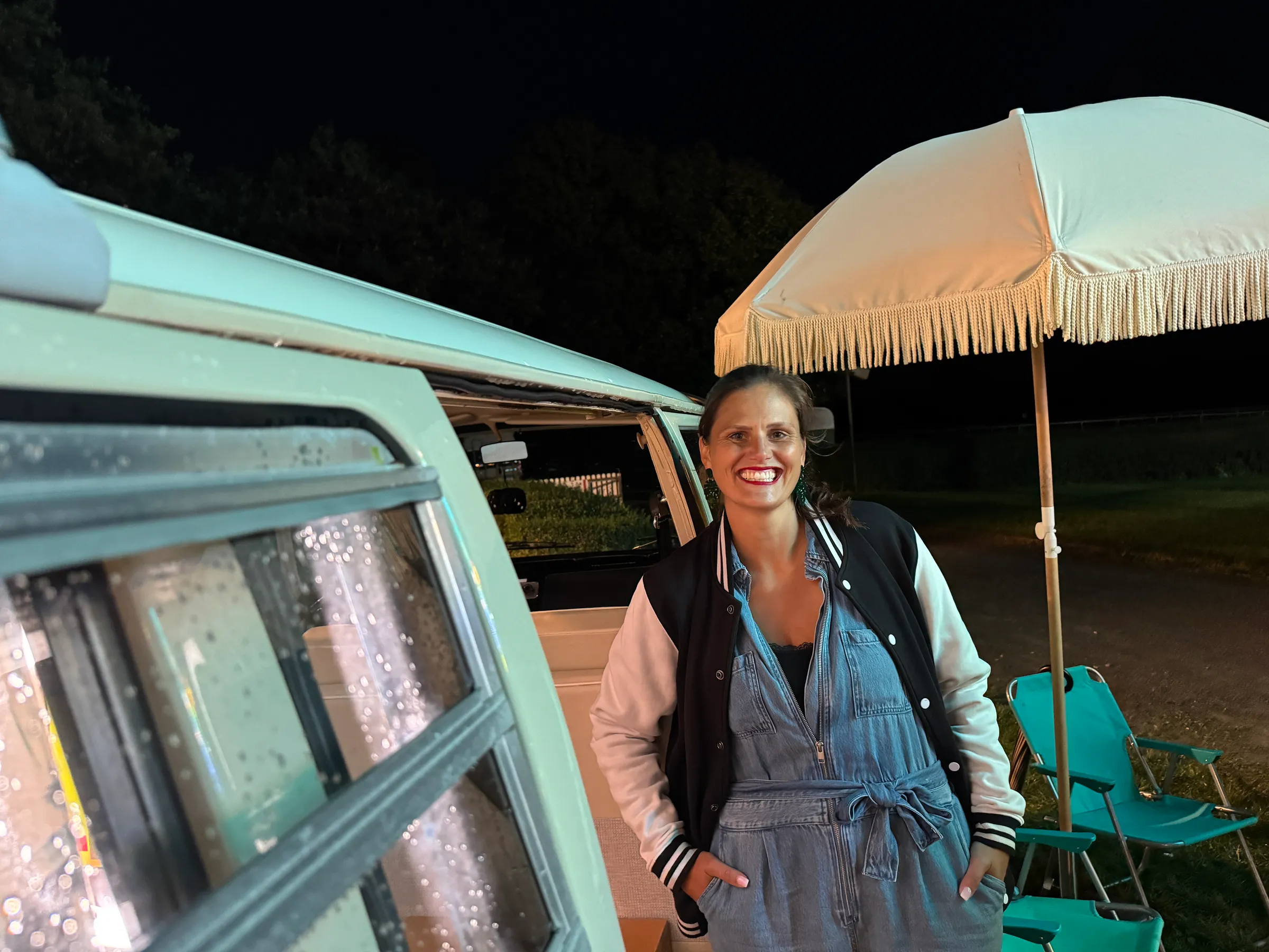 Smiling host in denim dungarees and a varsity jacket standing next to the photo booth bus at night, with a fringed parasol and folding chairs behind her