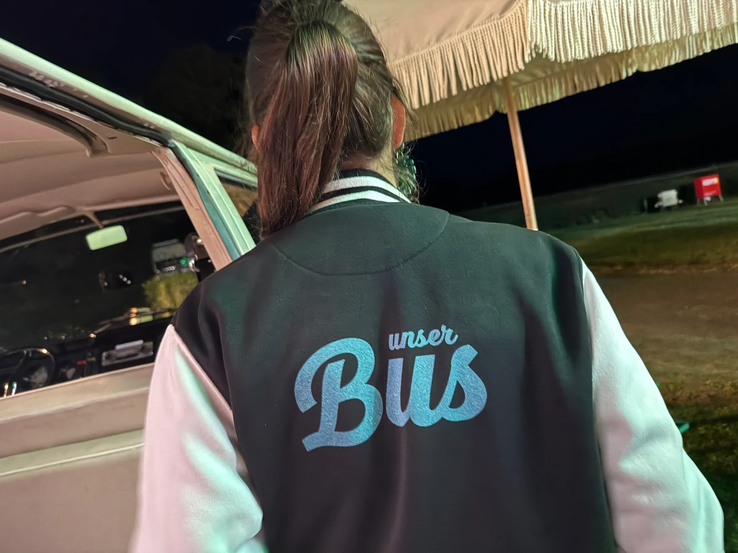 Host in a college jacket with the lettering ‘unser Bus’ standing in the evening next to the open photo booth bus under a fringed umbrella