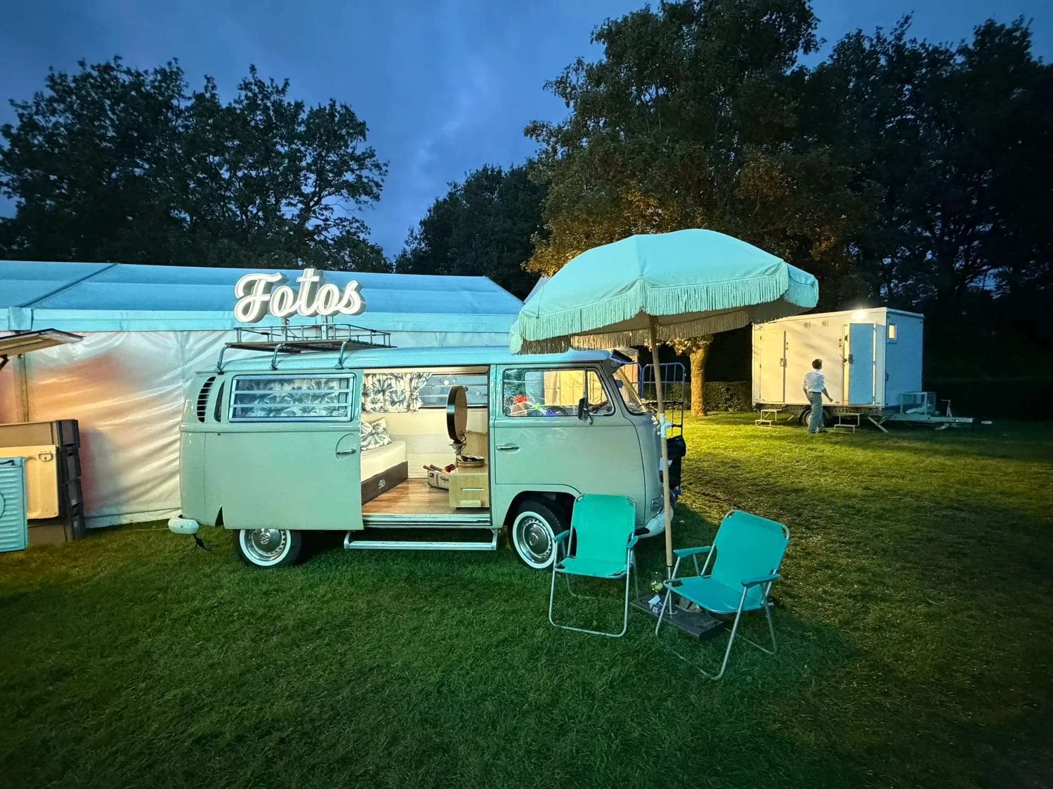 Mint-green photo booth bus with a neon ‘Fotos’ sign, a fringed parasol and two camping chairs on a meadow at dusk